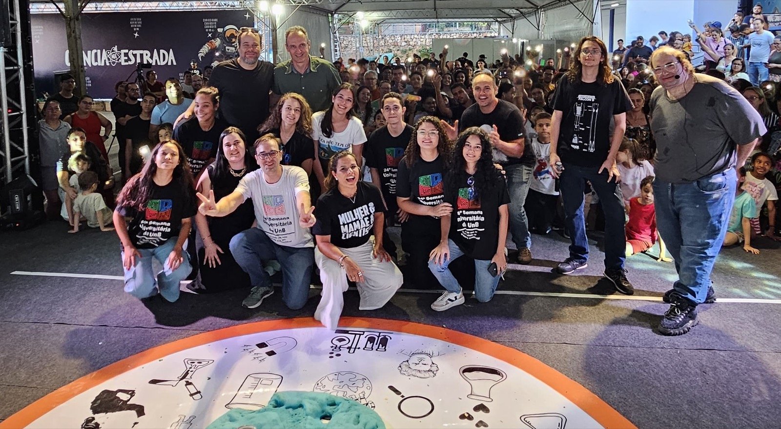 Equipe e público reunidos em frente ao palco da Ciência na Estrada, com banner do projeto ao fundo
